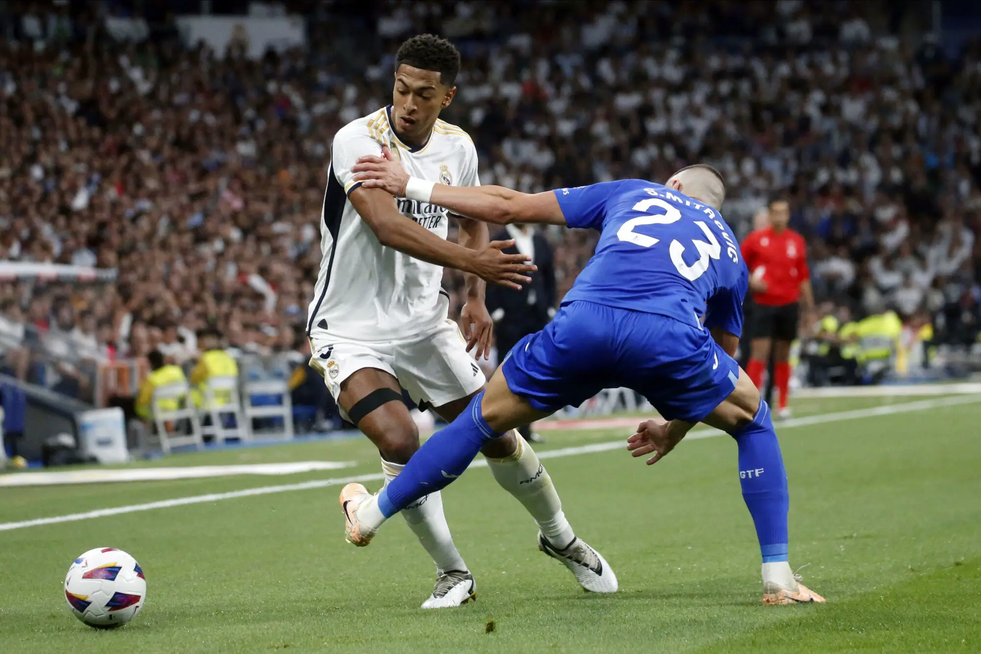 Image de Football. Dans un match palpitant au Santiago Bernabéu, le Real Madrid a décroché une victoire spectaculaire de 2-1 contre Getafe lors de la 4e journée de la Liga. Les Merengues, dirigés par Carlo Ancelotti, ont affronté une équipe de Getafe résistante qui cherchait à créer la surprise. Malgré l'absence de joueurs clés tels que Vinicius Junior et Thibaut Courtois, le Real Madrid a pris le contrôle du match mais a concédé un but tôt en raison d'une erreur défensive. Borja Mayoral a profité d'une confusion en défense pour donner l'avantage à Getafe. Le Real Madrid a égalisé en début de seconde période grâce à Joselu, qui a converti une passe de Modric. La dynamique s'est ensuite inversée en faveur du Real, avec Kroos qui a touché le poteau. Cependant, c'est Jude Bellingham qui a volé la vedette, profitant d'un rebond en temps additionnel pour assurer la victoire au Real Madrid. L'impact de Bellingham cette saison a été indéniable, et ses exploits de dernière minute ont maintenu le Real en tête du classement de la Liga. Que pensez-vous de ce match ?